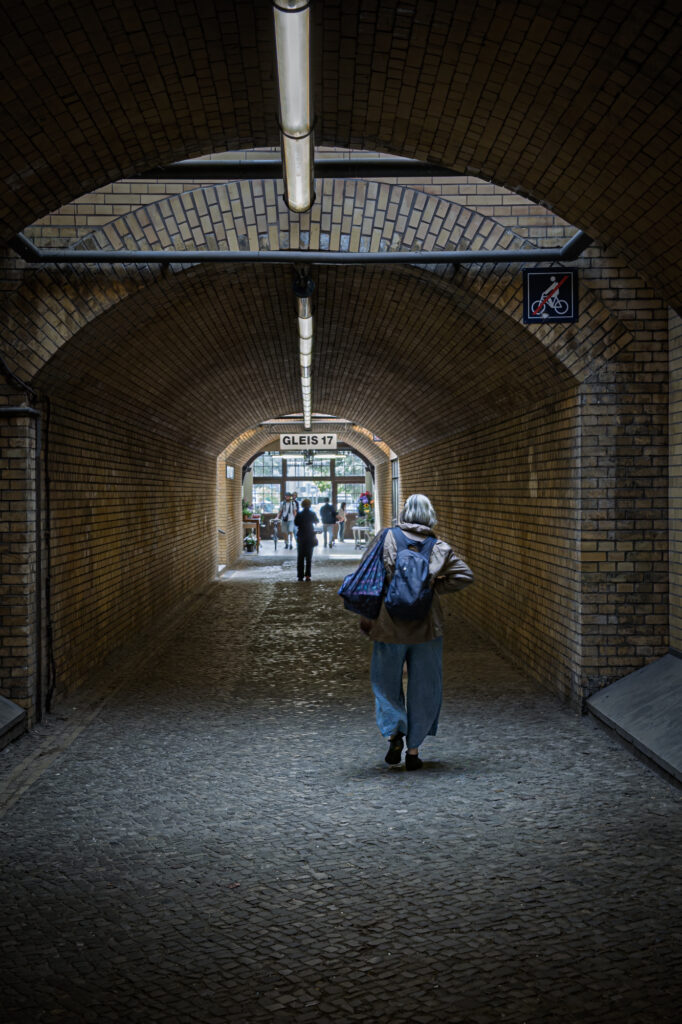 This photograph shows the brick underpass beneath Platform 17 at Grunewald station in Berlin. A single figure walks away from the camera, carrying a bag and wearing a backpack. The arched ceiling and repeating brickwork create strong symmetry. Overhead lights run along the centre line, guiding the viewer&rsquo;s gaze toward the bright opening at the far end.

A sign reading Gleis 17 hangs above the exit. Today, commuters and visitors pass through this space without pause. During the Second World War, Jews were forced through these platforms and loaded onto trains bound for concentration camps. This underpass formed part of that route.

The image holds tension between past and present. The bricks remain. The structure remains. Daily life continues. Yet the location carries the memory of deportation and loss. The solitary figure becomes a quiet witness, walking through a corridor shaped by history.