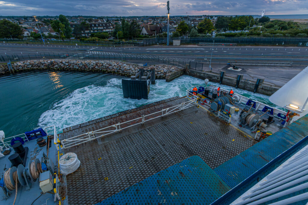 The Mont St Michel ferry eases into Ouistreham under a bruised evening sky, its stern ramp glistening with salt and wear. The whir of winches and churn of propellers echo against the harbour wall, marking the end of a cross-channel journey. Shipworkers in orange, watchful silhouettes, stand poised, custodians of motion between land and sea. Beyond the harbour, the quiet town waits under the last blush of daylight, its streets still, its horizon crowned by a distant wheel turning gently above the beach.