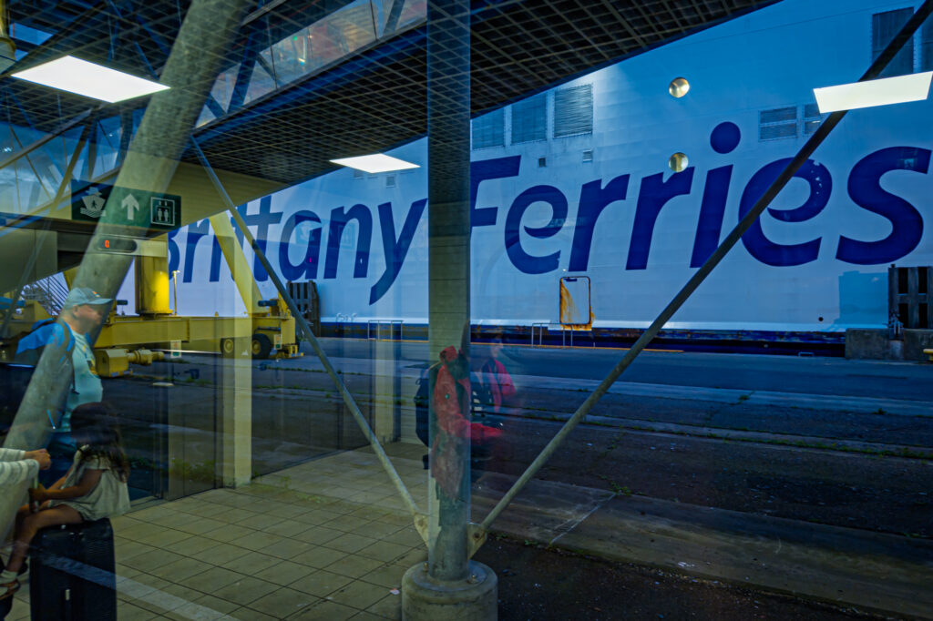 Passengers wait within the glass corridor of the Ouistreham ferry terminal as the vast hull of Brittany Ferries Mont St Michel looms outside. The scene becomes a layered reflection of the journey and pause, with travellers&rsquo; silhouettes merging with the industrial geometry of beams, lights, and the ship&rsquo;s colossal lettering. Through the transparent barriers, reality and memory overlap: the promise of landfall against the lingering rhythm of the sea.