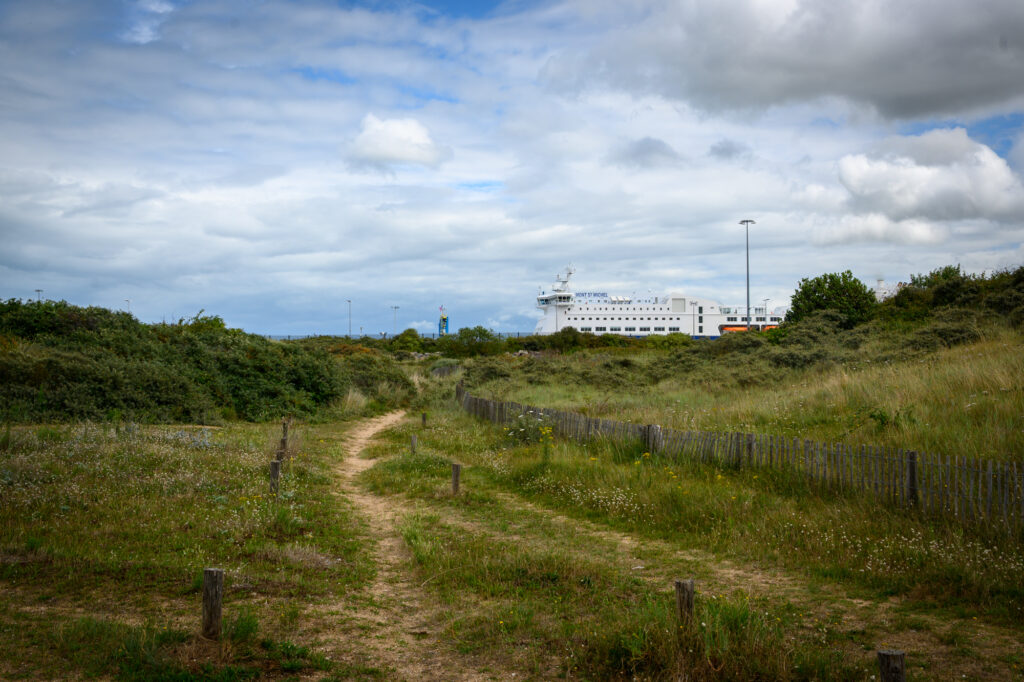 A winding coastal path leads through the wild grasslands of Ouistreham&rsquo;s dunes, guiding the eye toward the towering Brittany Ferries vessel Mont St Michel. The white ship, poised for departure, stands out sharply against the green earth and the grey-blue sky above. The ferry&rsquo;s presence introduces a human rhythm into this tranquil natural setting a moment caught between stillness and motion, land and sea, departure and return.