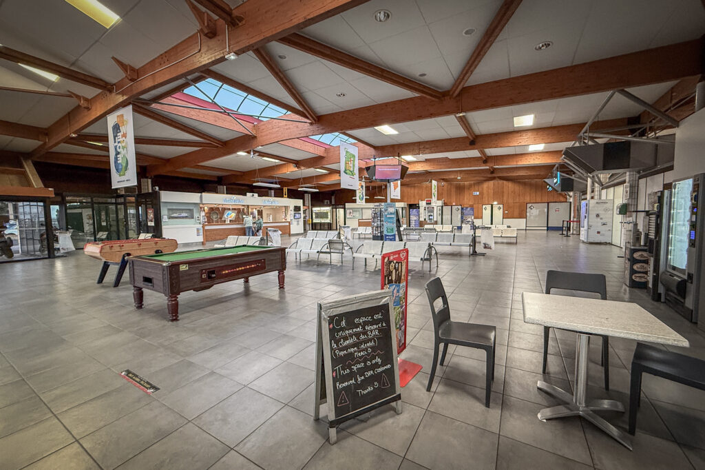 Inside the Ouistreham Ferry Port terminal in Normandy, France, light spills gently through the skylights, illuminating a calm moment between journeys. The hall&rsquo;s wooden beams and tiled floor frame an atmosphere of transience, where games of table football and pool stand ready to distract travellers from the slow rhythm of waiting. A chalkboard sign reserves a modest caf&eacute; space, a reminder of human order amid the ebb and flow of travel. Outside, ferries await their next crossing to England, while inside, time seems to pause, caught between departure and arrival.